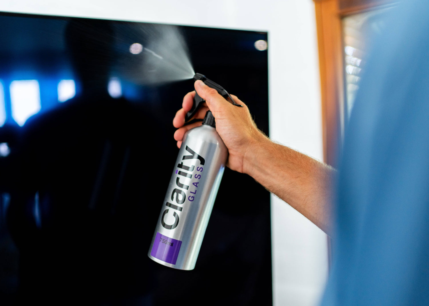 Spraying Clarity Glass Cleaner on TV screen - ammonia-free formula safely removes dust and fingerprints without damaging anti-glare coatings.